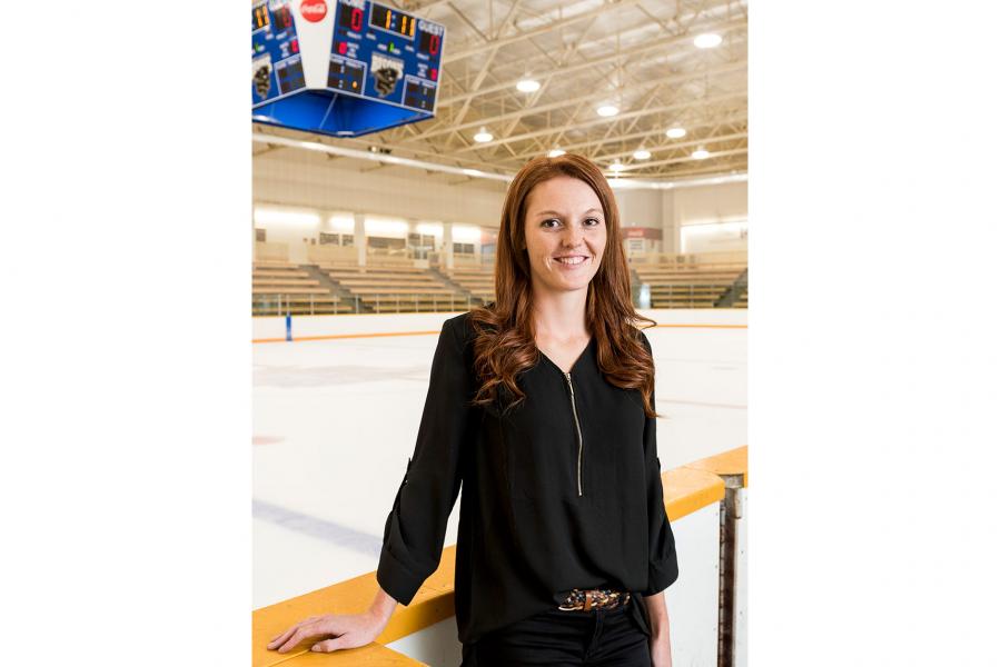 Caitlyn Fyten a smiling young Caucasian woman with red hair and freckles stands in front of the hockey ring boards with the rink and stands in the background.