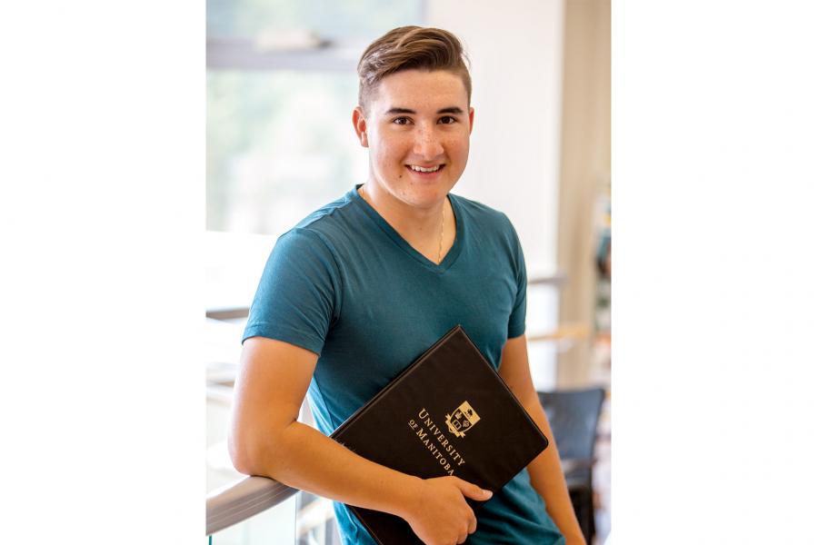 Riley Chartrand, a smiling young Metis male stands leaning against a railing with a University of Manitoba clipboard in hand in a light and airy space.