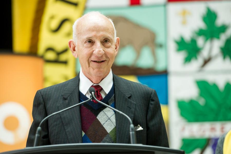 Ernest Rady, a senior Caucasian male in a suit, sweater vest and tie stands behind a podium with dual mics.