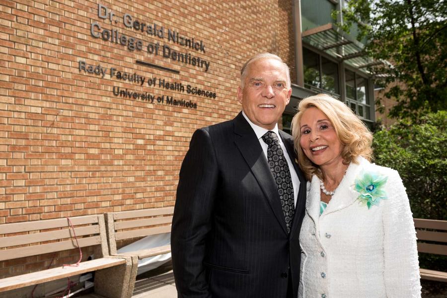 A senior Caucasian couple Dr, Gerald and Mrs. Ressa Niznick, in business attire stand in front of an orange-red brick modern building. The sign on the building reads Dr. Gerald Niznick College of Dentistry.
