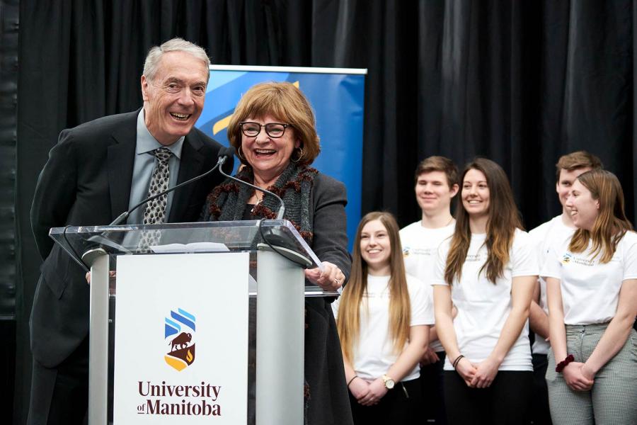 Dr. Gerry Price a senior Caucasian male and his wife Barb Price a senior Caucasian female stand in business attire behind a dual mic podium with a group of Engineering students smiling behind them on stage. 