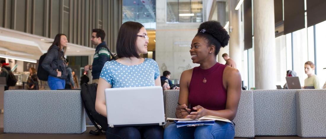Two students seated together at a bench, one has a laptop open on her lap while they talk.