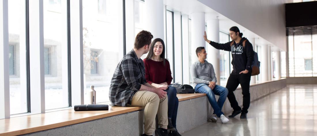 Four students sit on a bench in a brightly lit hallway lined with windows.