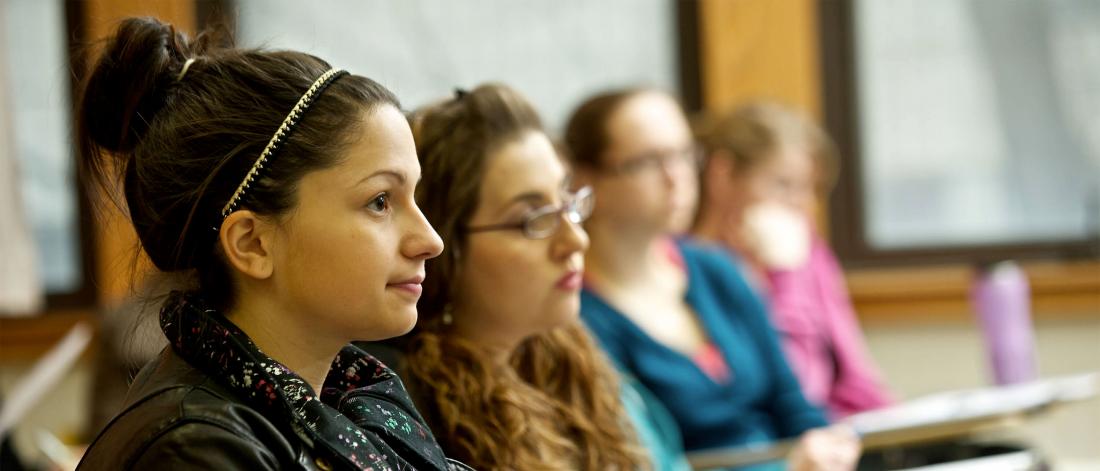 Close-up profile shot of a young woman sitting in a classroom, with three other women sitting in the rows beside her.