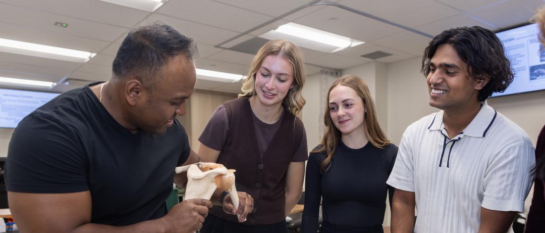 A group of four students gathered around a male instructor, demonstrating a bone model in a lab setting.