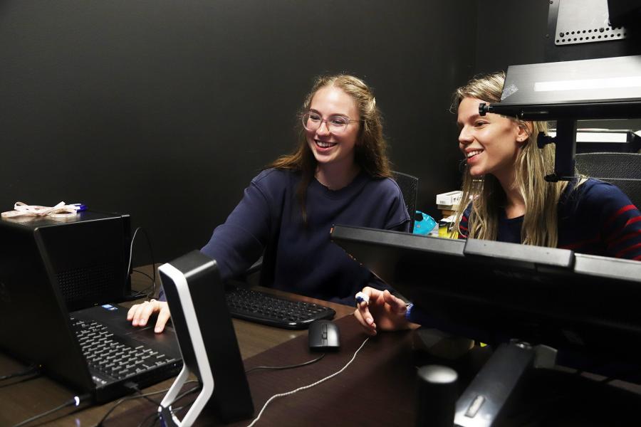 Two students sit in a lab setting in front of a laptop with one student wearing a small monitoring device on one finger.