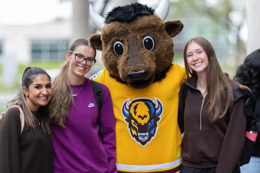 Three individuals stand on either side of the University of Manitoba mascot (Billy the Bison) in an outdoor setting.
