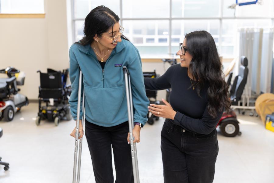 Two women in a rehabilitation lab. One uses crutches, while the other gives support.