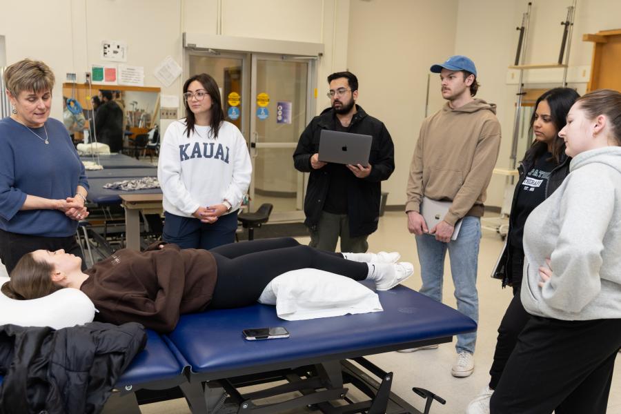 A group of six students is gathered around a therapy table in a clinical setting, listening to an instructor, while one person lies on the table.
