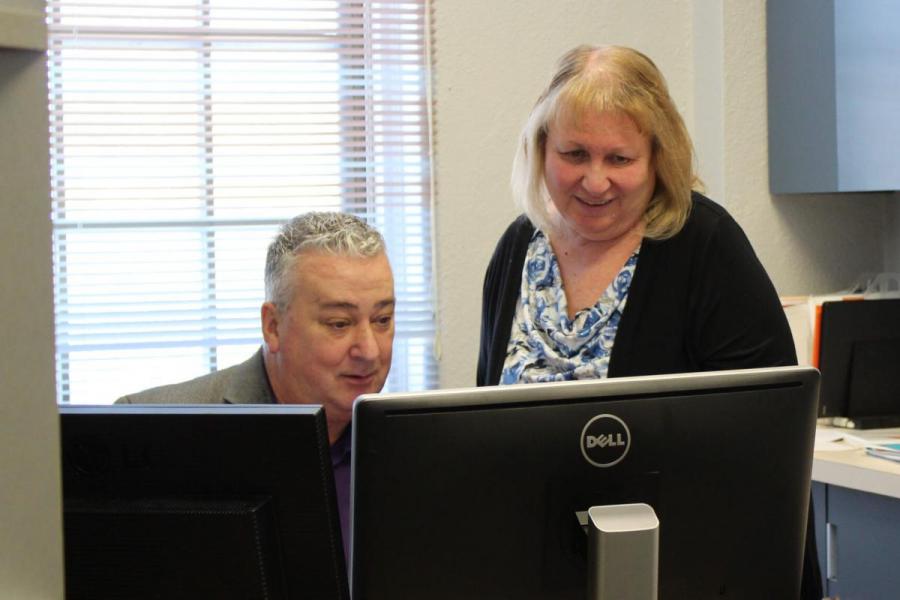 Man sitting down in a grey suit looking at two monitor screens, with a woman in a cardigan standing looking over his shoulder at the screen.