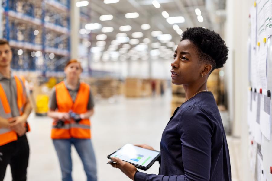 Woman holding a tablet in a warehouse managing other people with safety vests.
