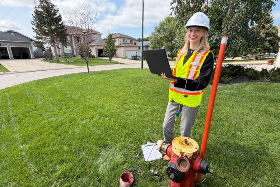 Student standing on the grass with a computer hooked up to a red and yellow fire hydrant. She is wearing a safety vest and hat.