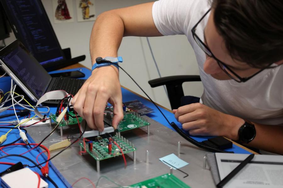 Student wearing a t-shirt and glasses sitting at a desk, hovering over a several circuits.