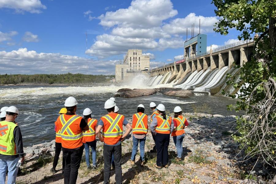 Group of people standing outside a hydro dam on a sunny day wearing safety vests and hats.