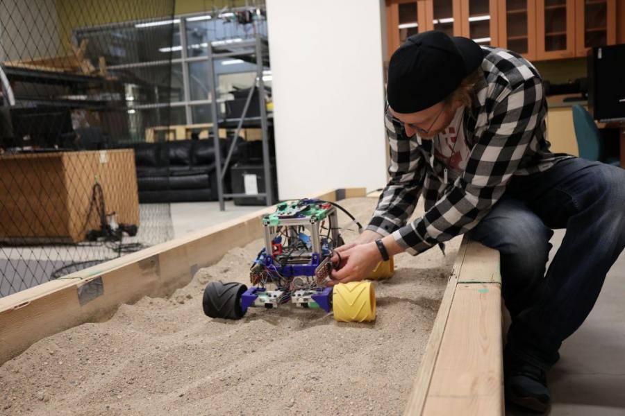 Student wearing plaid shirt testing a small mechanical vehicle in a test sand pit in a lab.