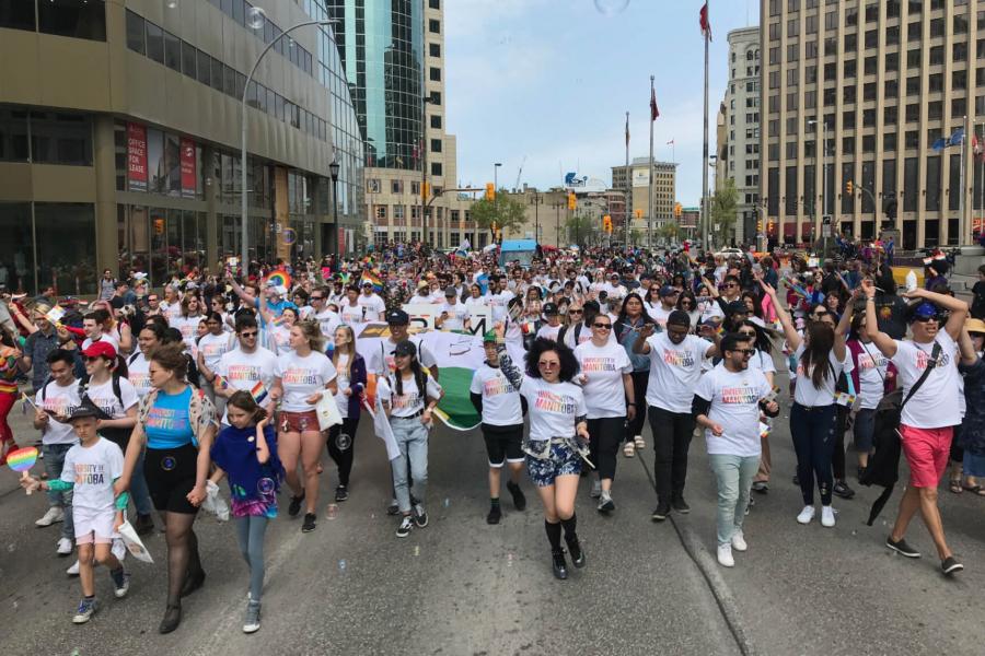 A large group of people march together on the street wearing matching white tshirts for Pride Day.