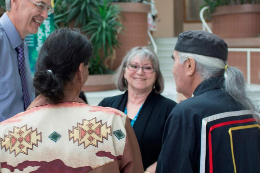 A group of people talking, two are dressed in traditional Indigenous clothing.