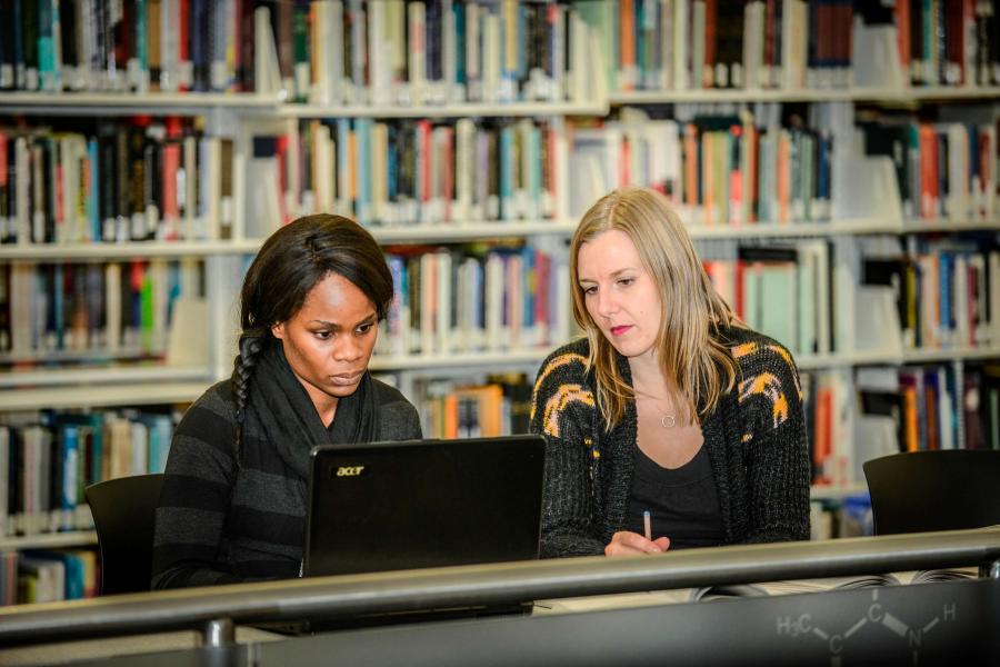 Two female students sitting side by side, looking at a laptop screen in a library.
