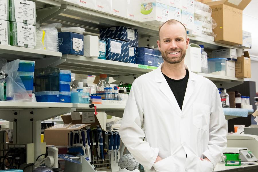 Man in a white lab coat stands in front of fully stocked lab shelves.