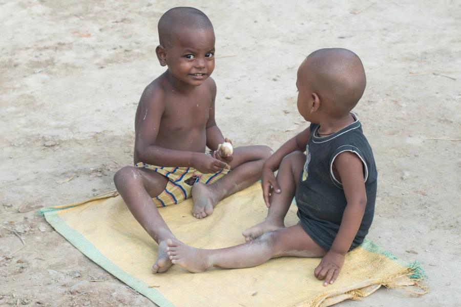 Two toddlers living in an impoverished community in India, sit outside, playing on a mat.