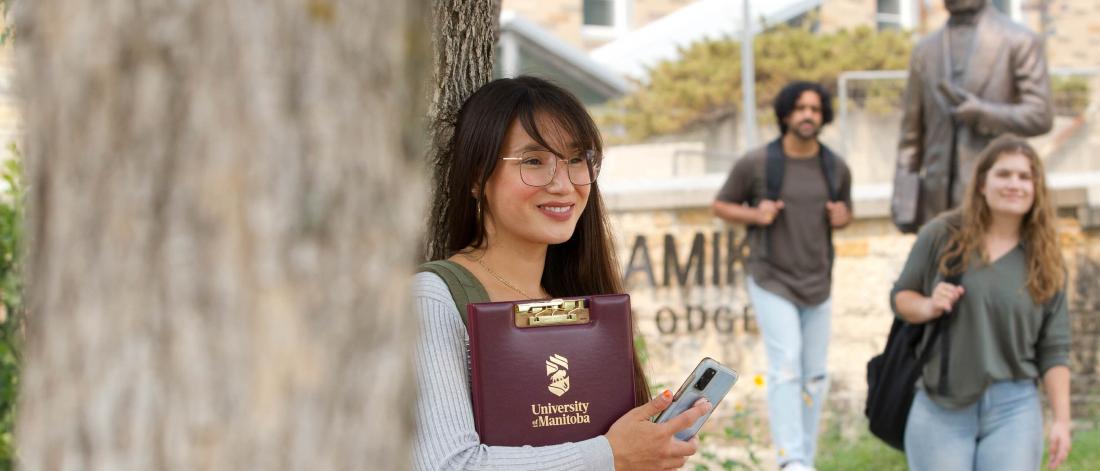A student standing outside Migizii Agamik leans against a tree holding a folder with a University of Manitoba logo on it. Two other students smile as they walk by. 