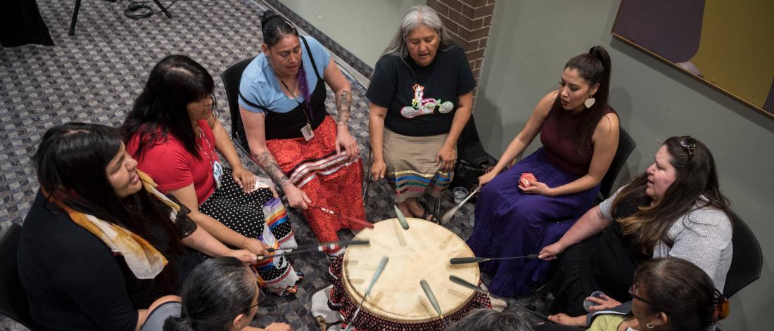 A group of women gathered in a circle beating a PowWow drum.