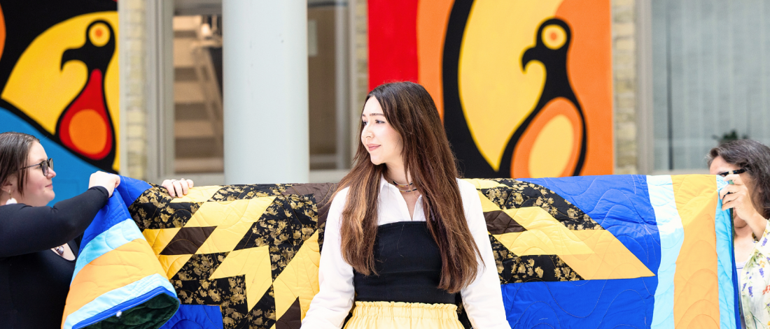 Three women hold up a colourful star blanket behind a woman wearing a yellow ribbon skirt during a blanketing ceremony.