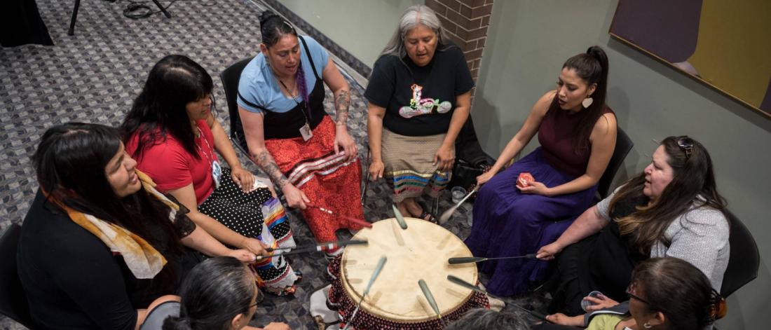 Participants sit in a circle, singing and drumming together during a cultural gathering.