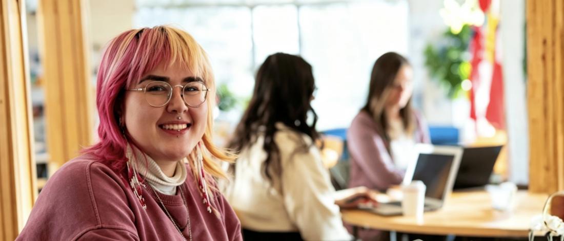 Smiling student with pink hair working on a tablet in a bright study space, with other students in the background.