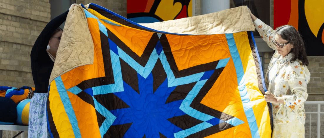 Two people hold up a brightly coloured star-pattern quilt during the Honouring the Indigenous Campus Community ceremony at the University of Manitoba.