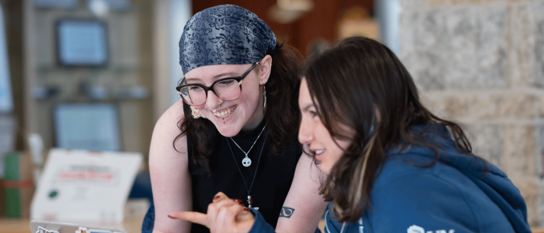 Two students smiling and looking at a laptop together in a campus common area.