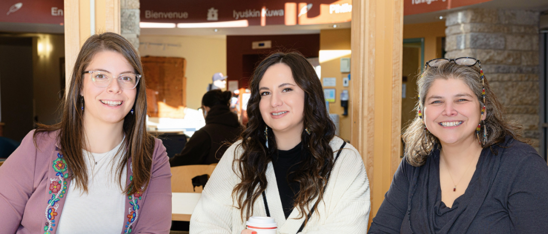 Three people smiling while seated together at a table in a bright campus common area.