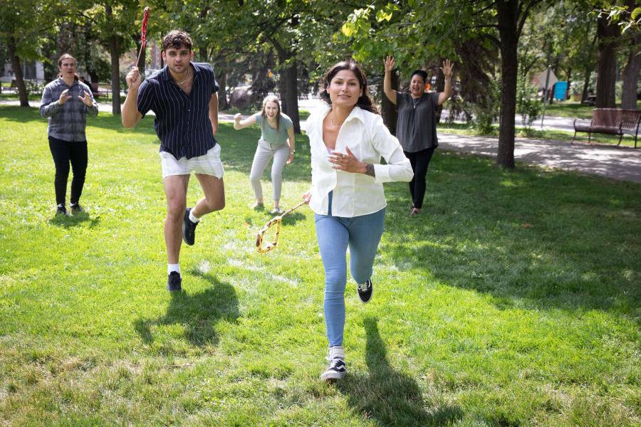 Two students running with stick in hand while 2 people cheer them on in the background.