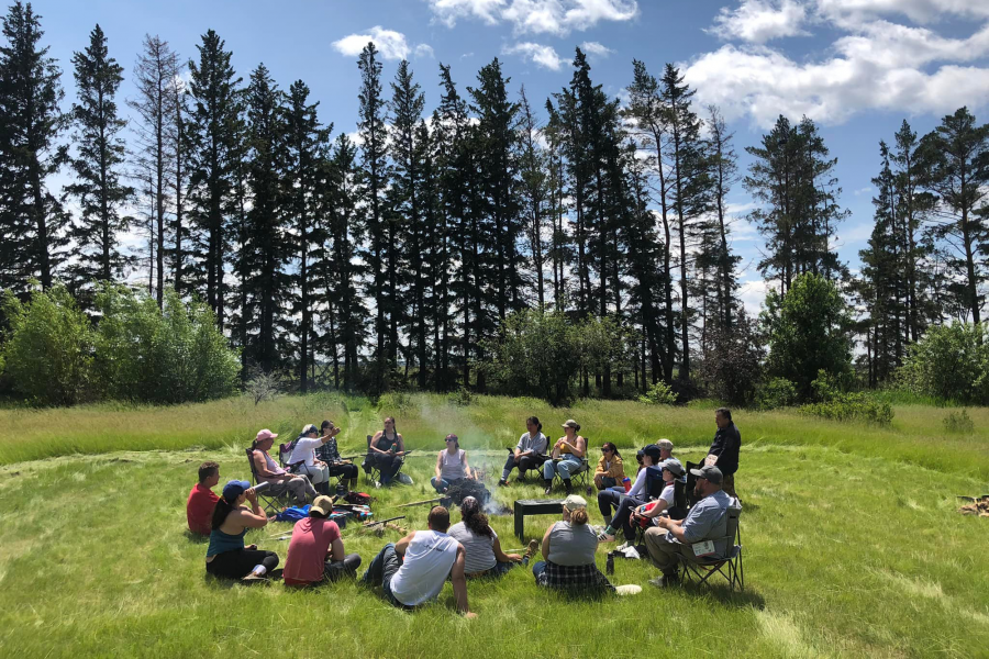 A group of students sitting on the grass in a circle with tall trees in the background