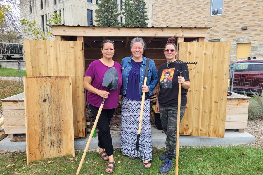 Three women, lodge caretakers, stand in front of a woodshed holding tools