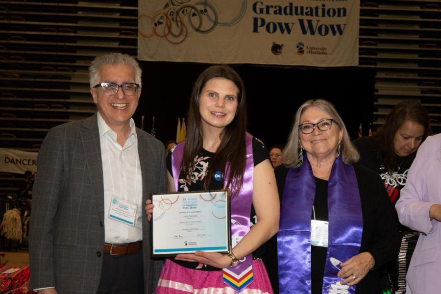 Student in a ribbon skirt and Indigenous graduation stole poses with graduation certificate at grad pow wow