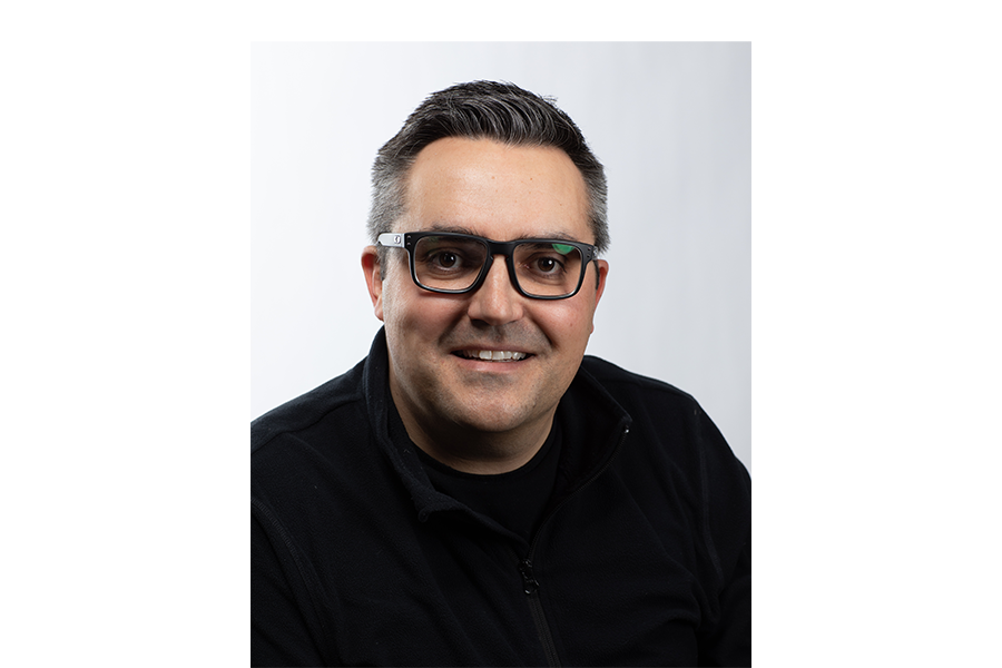 A man with short dark hair and glasses is smiling and wearing a black jacket against a plain white background.