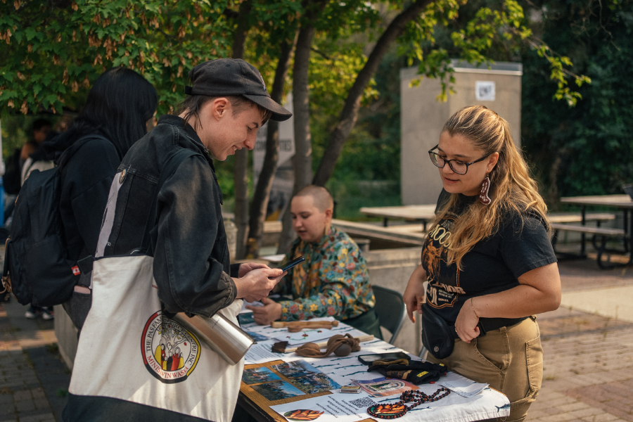At an outdoor booth, a woman wearing earrings speaks with a student, with cultural items and promotional materials displayed on the table.