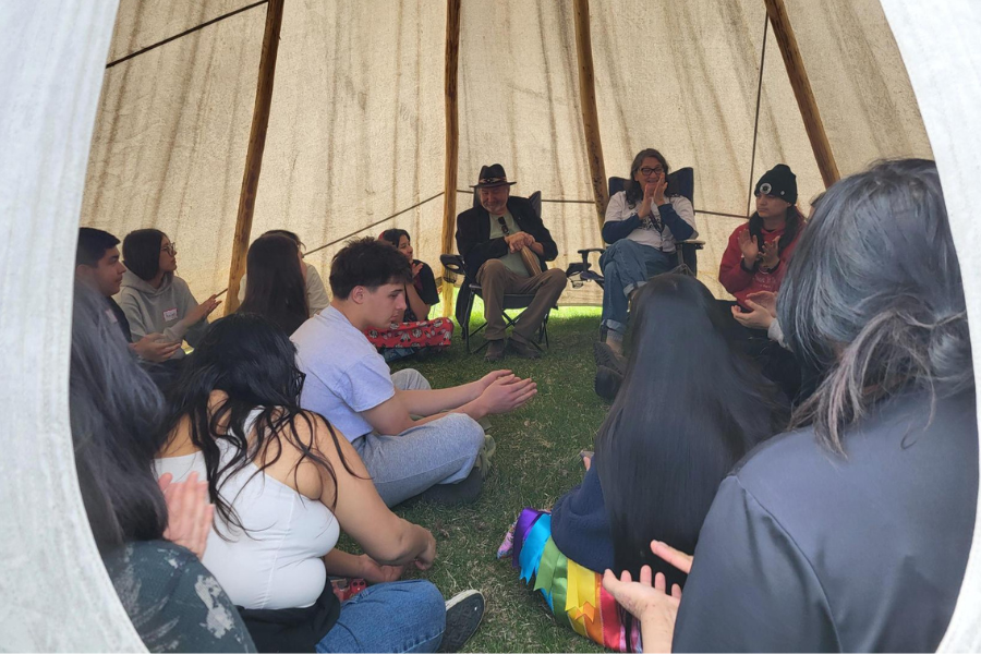 Inside a tipi, Elders speak while students sit in a circle listening and clapping.