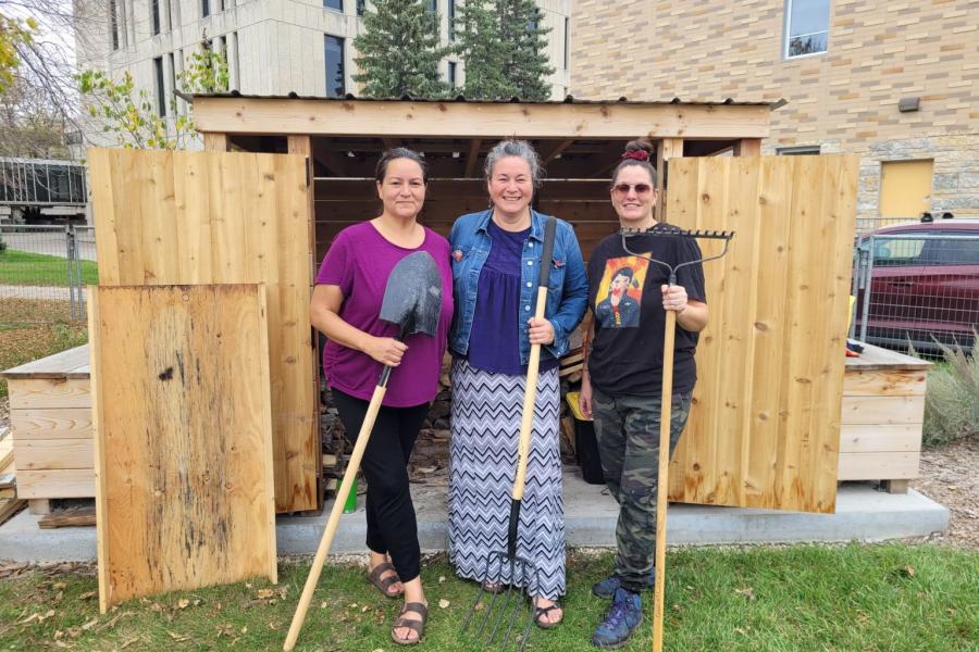Three women standing in front of a wooden garden shed, smiling and holding gardening tools — a shovel, pitchfork, and rake — on a grassy area at the University of Manitoba campus.