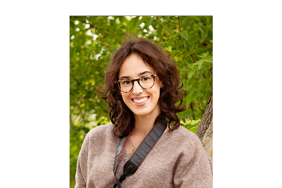 A woman in glasses standing in from of greenery looking at the camera.