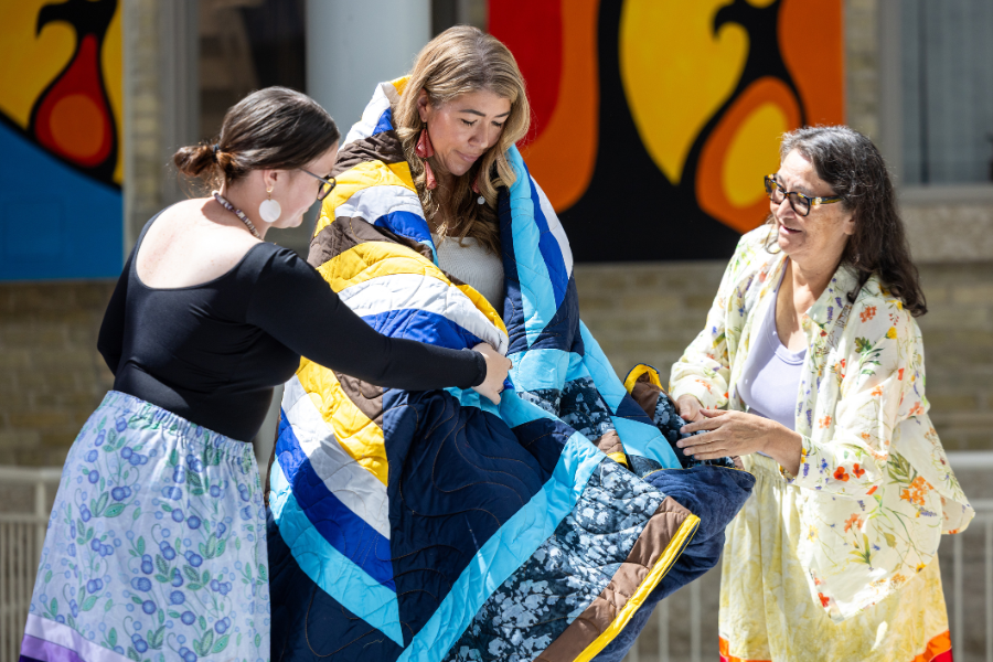 Elder and young women gently wrap a woman in a star blanket during the ceremony.