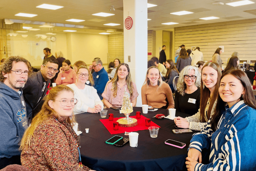 A group of people seated around a table, smiling during a community gathering indoors.