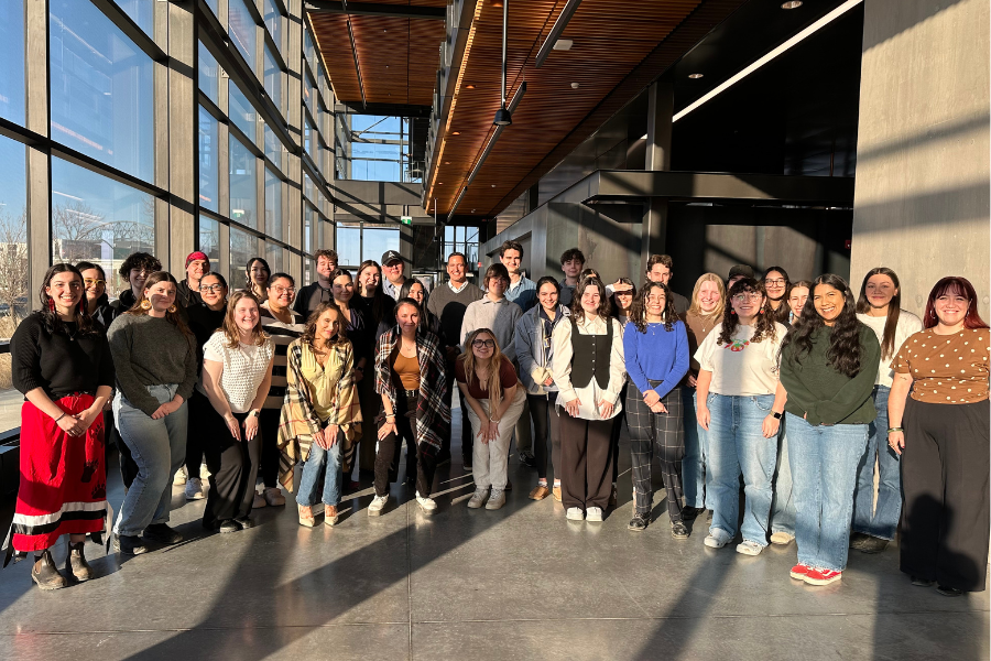 Group photo of students, faculty and staff gathered indoors in a bright campus space with floor-to-ceiling windows.