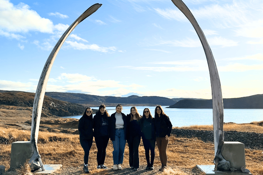 Five people standing beneath a large whale jawbone arch overlooking a coastal landscape and water.
