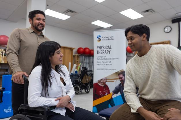 Two men talking to a woman sitting in a wheelchair standing in front of a physical therapy banner