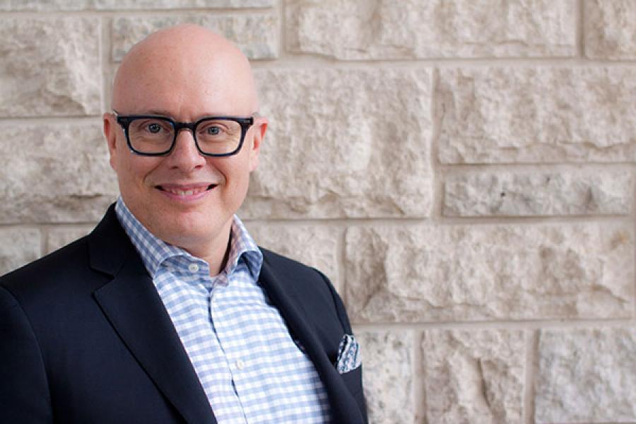 a bald man in suit jacket and pale blue checked shirt smiles at the camera in front of a pale tyndall stone wall
