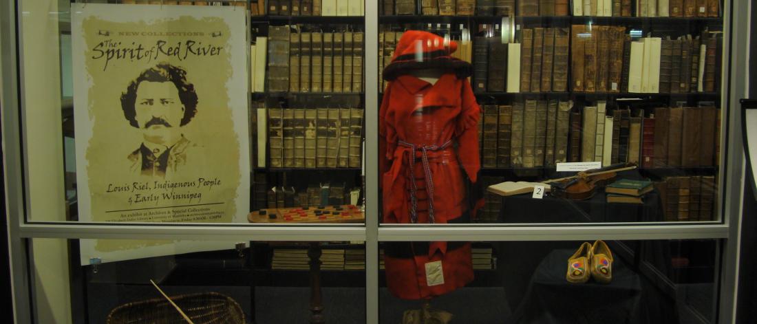The window at the entrance to the Archives showing an exhibit about the Red River Colony. There is a poster with a drawing of Louis Riel and a mannequin with a traditional red outfit.