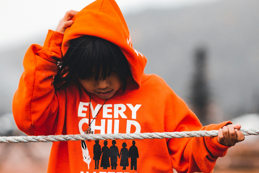 Indigenous child with black hair wearing an orange "every child matters" hoodie, with the hood up over their head. The child is outdoors in their community, looking down at the ground, one hand adjusting their hood, the other hand holding onto a rope that appears to be acting as a fence. 