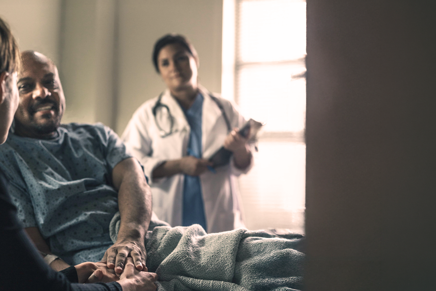 A man in a hospital bed and gown holds hands with his partner, looking at her optimistically, while a Female Physician in a white coat, wearing a stethescope, and holding a clipboard looks on from the background. 
